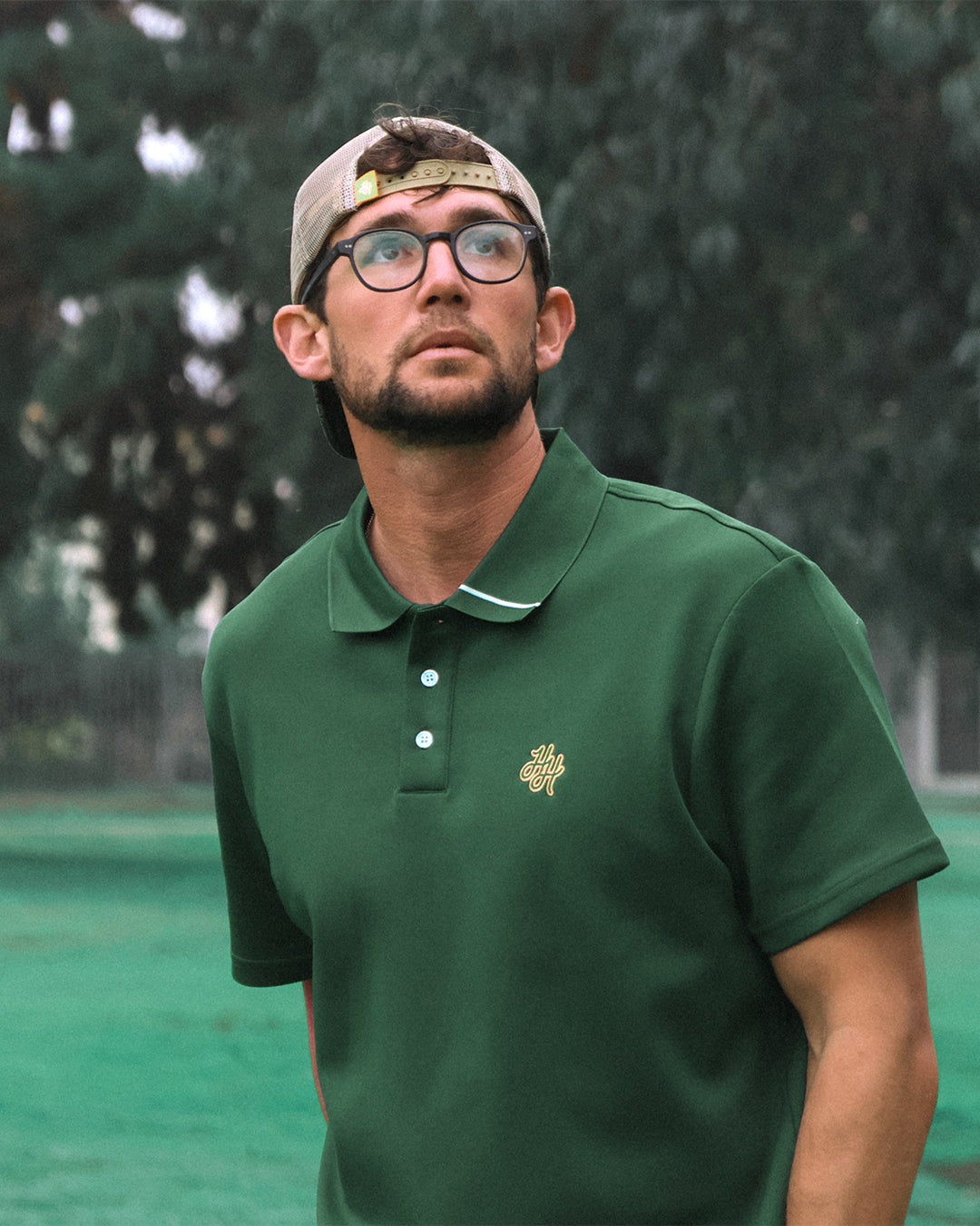 Man wearing a green polo shirt with a logo, standing outdoors with trees in the background.