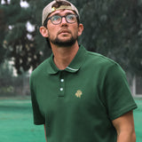 Man wearing a green polo shirt with a logo, standing outdoors with trees in the background.