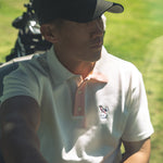 Man in a white polo shirt with a logo, sitting on a golf course.