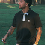 Man wearing a black polo shirt with a logo, standing outdoors on grass.