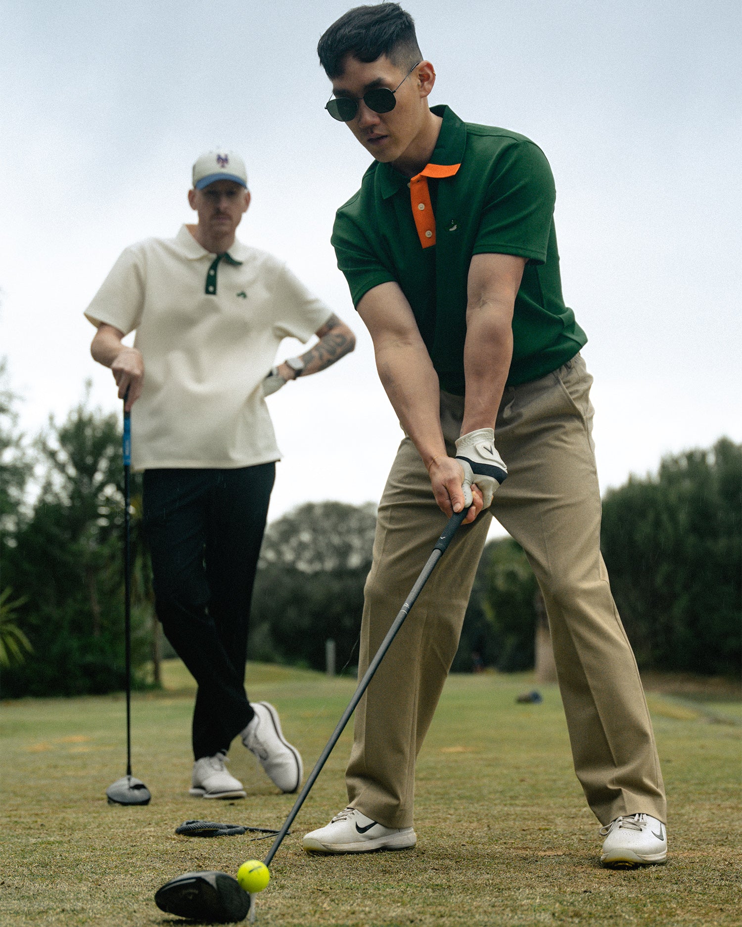 Two man standing holding golf clubs. One is wearing a green polo with orange accents the other is wearing a white polo with green accents.