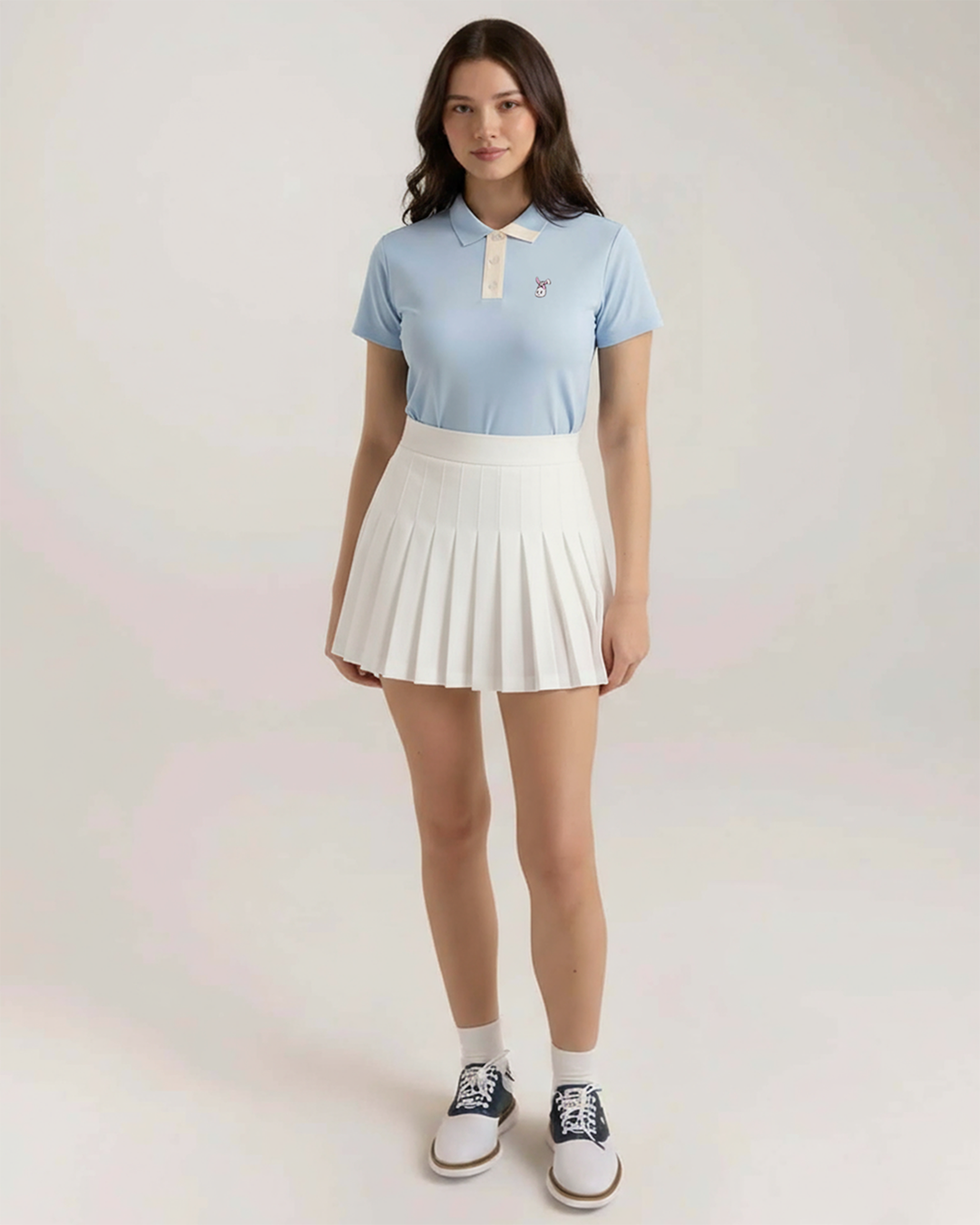 Woman wearing a light blue polo shirt and white pleated skirt on a plain background